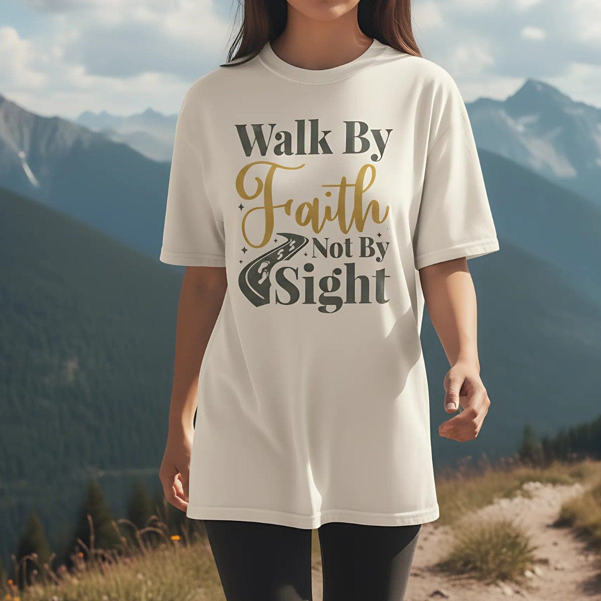 Christian woman wearing a Comfort Colors Christian t-shirt with the phrase 'Walk By Faith Not By Sight' in gold and gray lettering, styled with a road and footprints graphic, photographed outdoors on a mountain trail