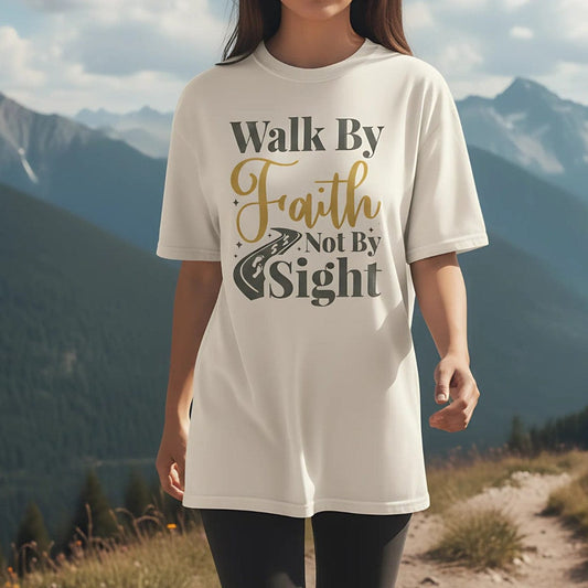 Christian woman wearing a Comfort Colors Christian t-shirt with the phrase 'Walk By Faith Not By Sight' in gold and gray lettering, styled with a road and footprints graphic, photographed outdoors on a mountain trail