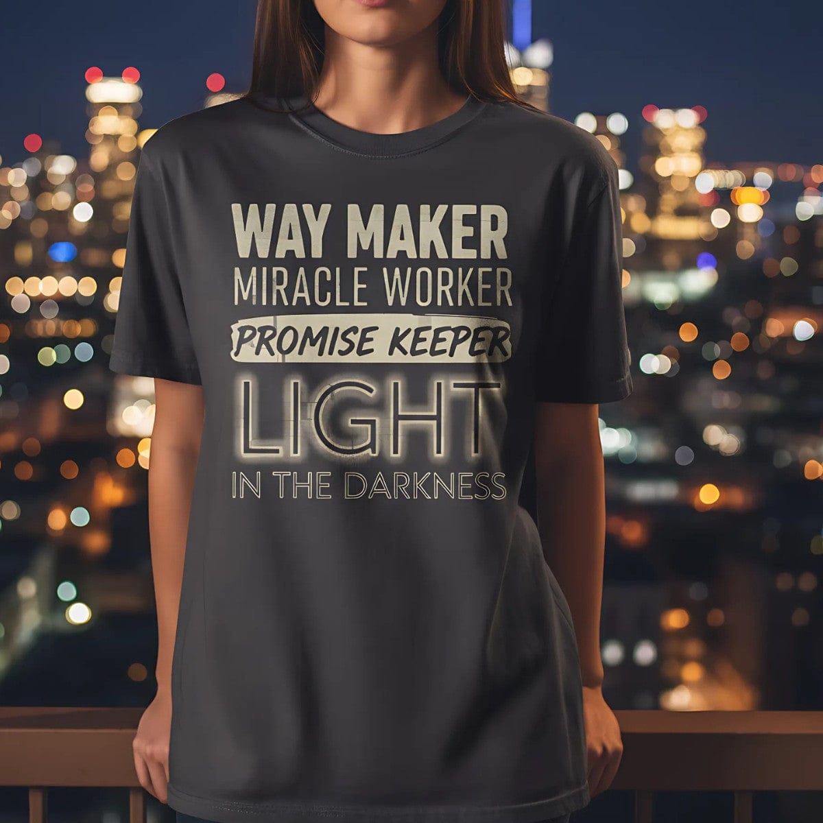Comfort Colors Christian t-shirt in charcoal grey worn by a woman in an urban night setting. Shirt features the worship phrase 'Way Maker, Miracle Worker, Promise Keeper, Light in the Darkness' in a glowing typography style.
