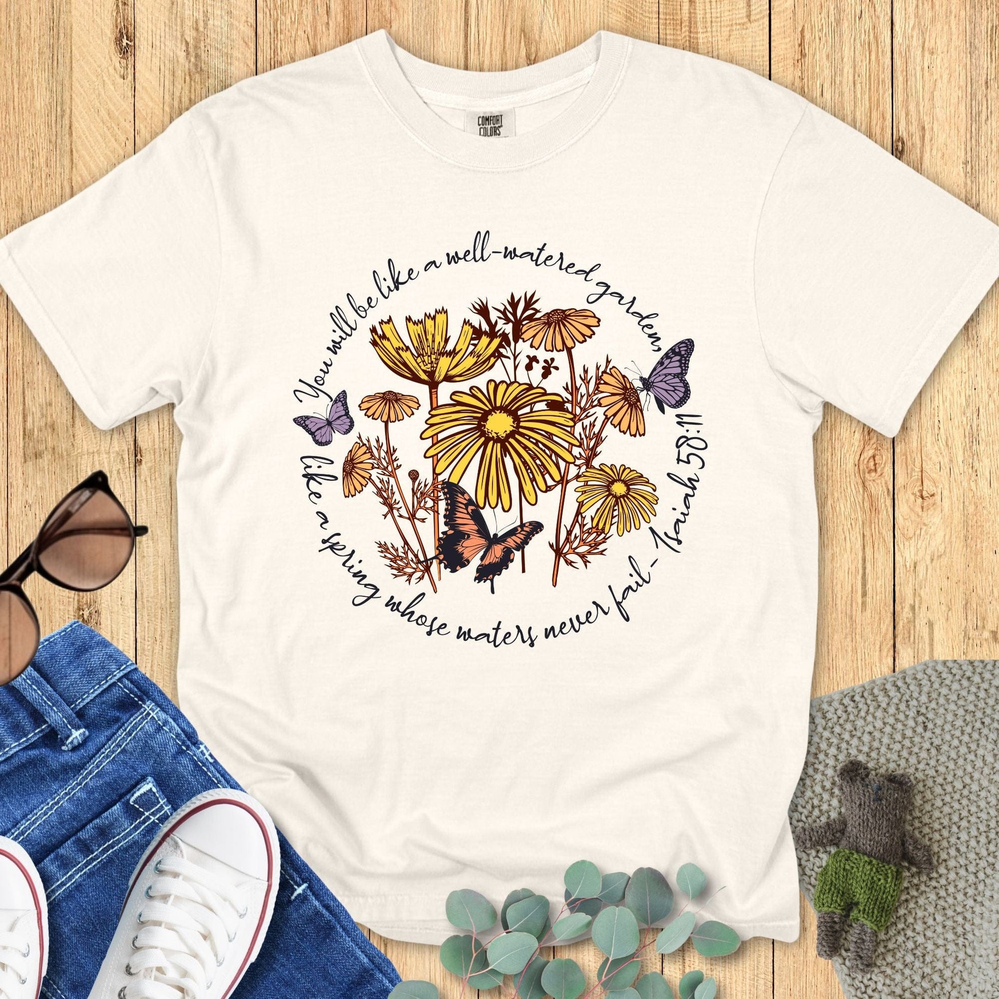 Christian Comfort Colors 1717 T-shirt in ivory with floral design featuring yellow daisies and butterflies. Verse reads: "You will be like a well-watered garden, like a spring whose waters never fail. Isaiah 58:11." Styled with denim jeans, sunglasses, and greenery on a wooden background.