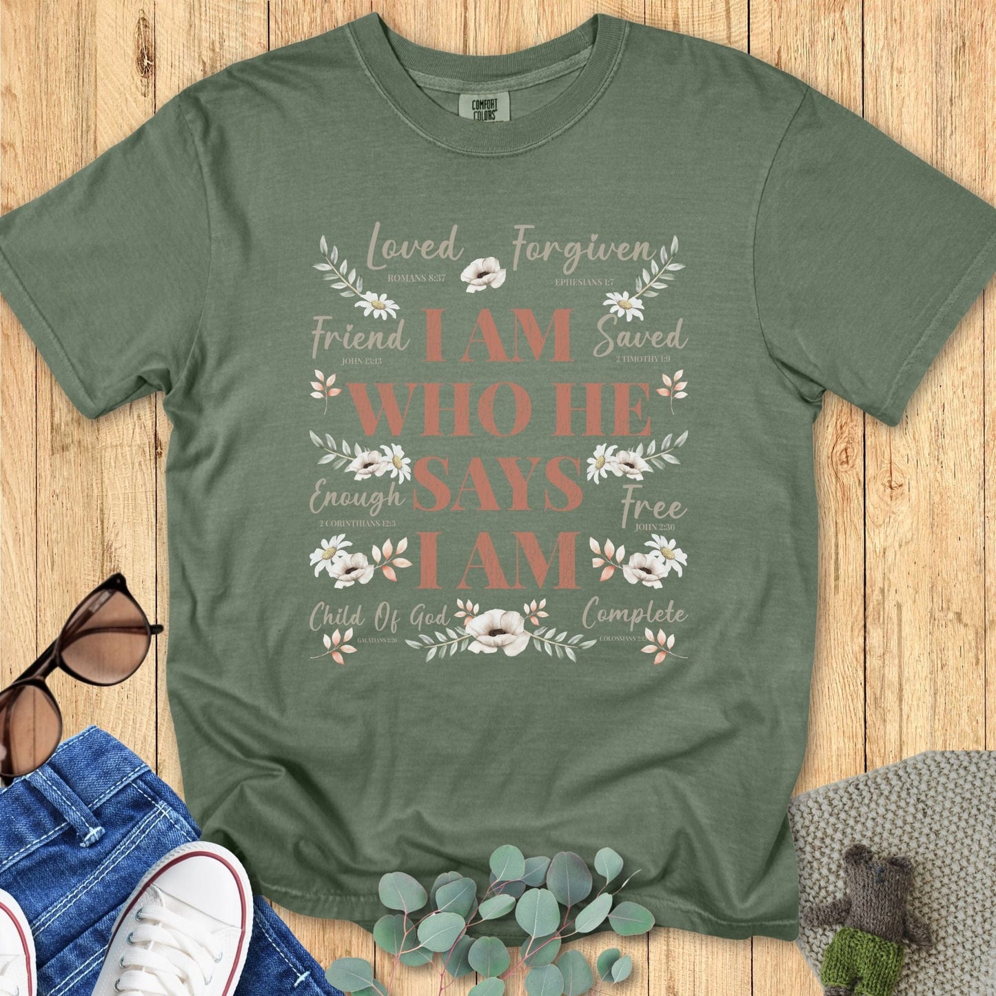 Sage green Christian women’s t-shirt featuring the affirmation “I Am Who He Says I Am” with scripture references and soft floral artwork, flat lay styled with jeans, sneakers, sunglasses, and eucalyptus for a boutique aesthetic