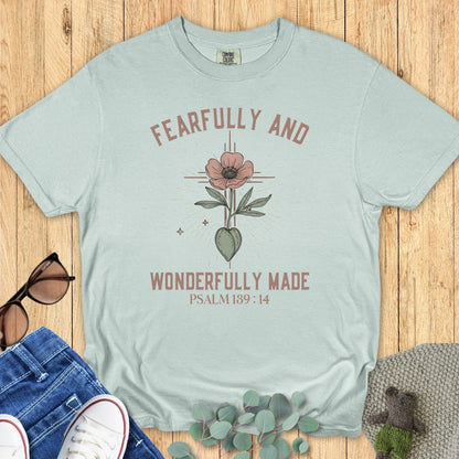 Flatlay of a Comfort Colors 1717 bay green “Fearfully and Wonderfully Made – Psalm 139:14” t-shirt with floral illustration, styled with jeans and sunglasses. A soft design from faith-based apparel for women collection.