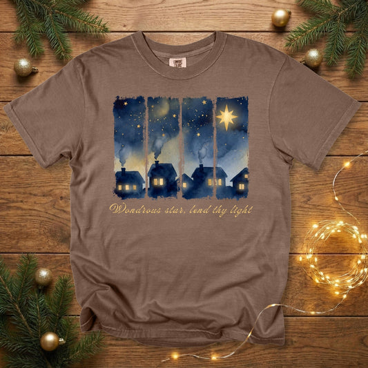 Comfort Colors 1717 espresso brown Christian women tshirt showing a watercolor village at night, illuminated homes, and a radiant Christmas star with the words “Wondrous star, lend thy light.” Cozy US faith wear tee styled on a wooden backdrop with festive greenery and soft holiday lighting.