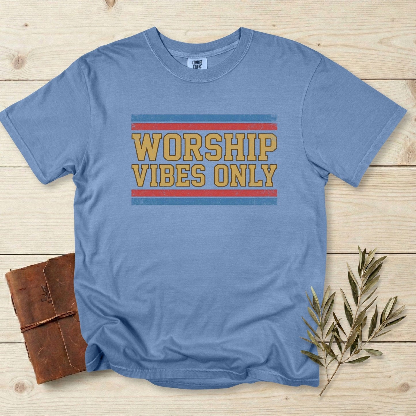 Comfort Colors 1717 blue jean Christian women tshirt featuring a bold retro “Worship Vibes Only” graphic in mustard, red, and blue stripes. Trendy US faith wear tee styled on a light wooden surface with a journal and olive branch.