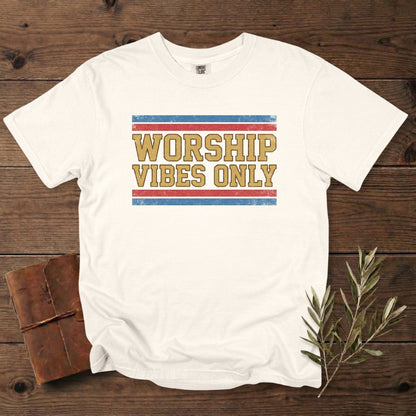 Comfort Colors 1717 ivory Christian women tee with a vintage “Worship Vibes Only” text graphic in mustard, red, and blue. Modern Christian tshirt for US faith wear, laid out on a rustic wooden background with journal and olive leaves.