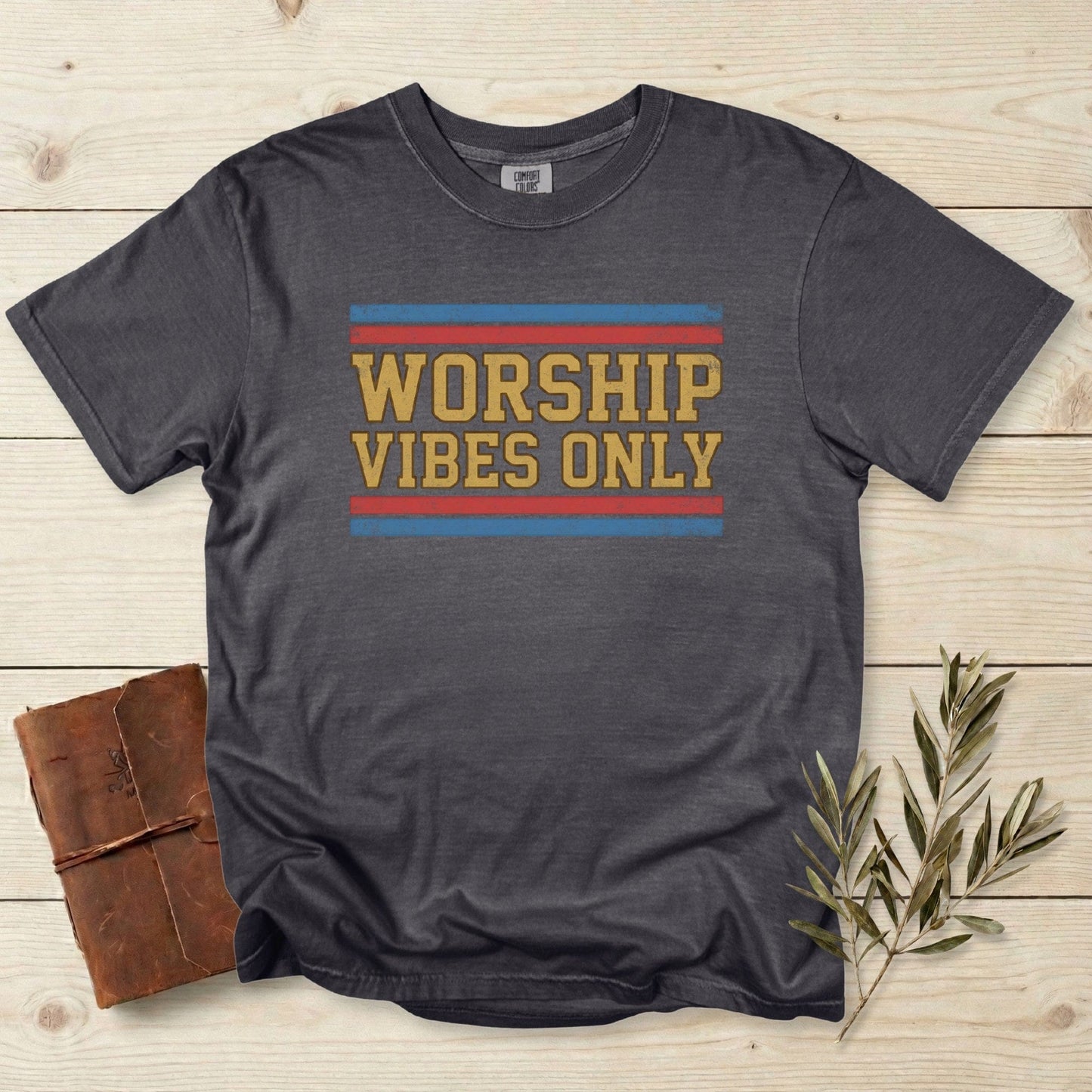 Comfort Colors 1717 pepper Christian women tee with a retro “Worship Vibes Only” design in mustard, red, and blue. Vintage-style Christian tshirt for US faith wear, displayed on a light wood background with a leather journal and olive branch.