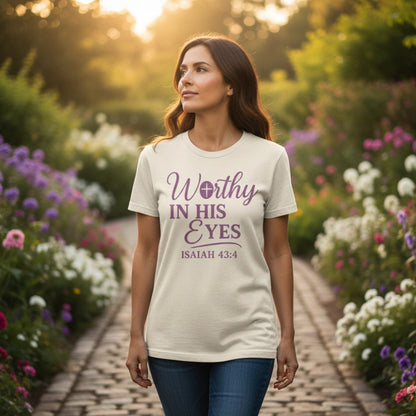 Christian women’s ivory t-shirt with 'Worthy in His Eyes – Isaiah 43:4' in purple text, worn by a woman walking through a vibrant flower garden at sunset, representing faith and encouragement.
