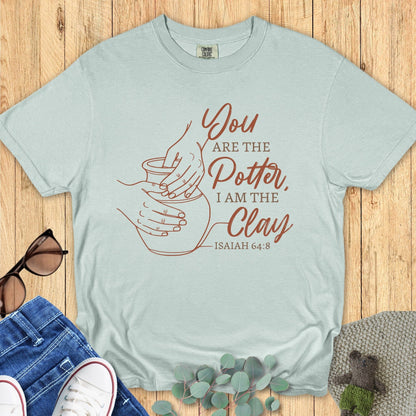 Comfort Colors Christian t-shirt in Bay color featuring pottery hands illustration with Isaiah 64:8 “You are the Potter, I am the Clay.” Christian apparel flat lay with jeans, shoes, and eucalyptus leaves on rustic wood background.