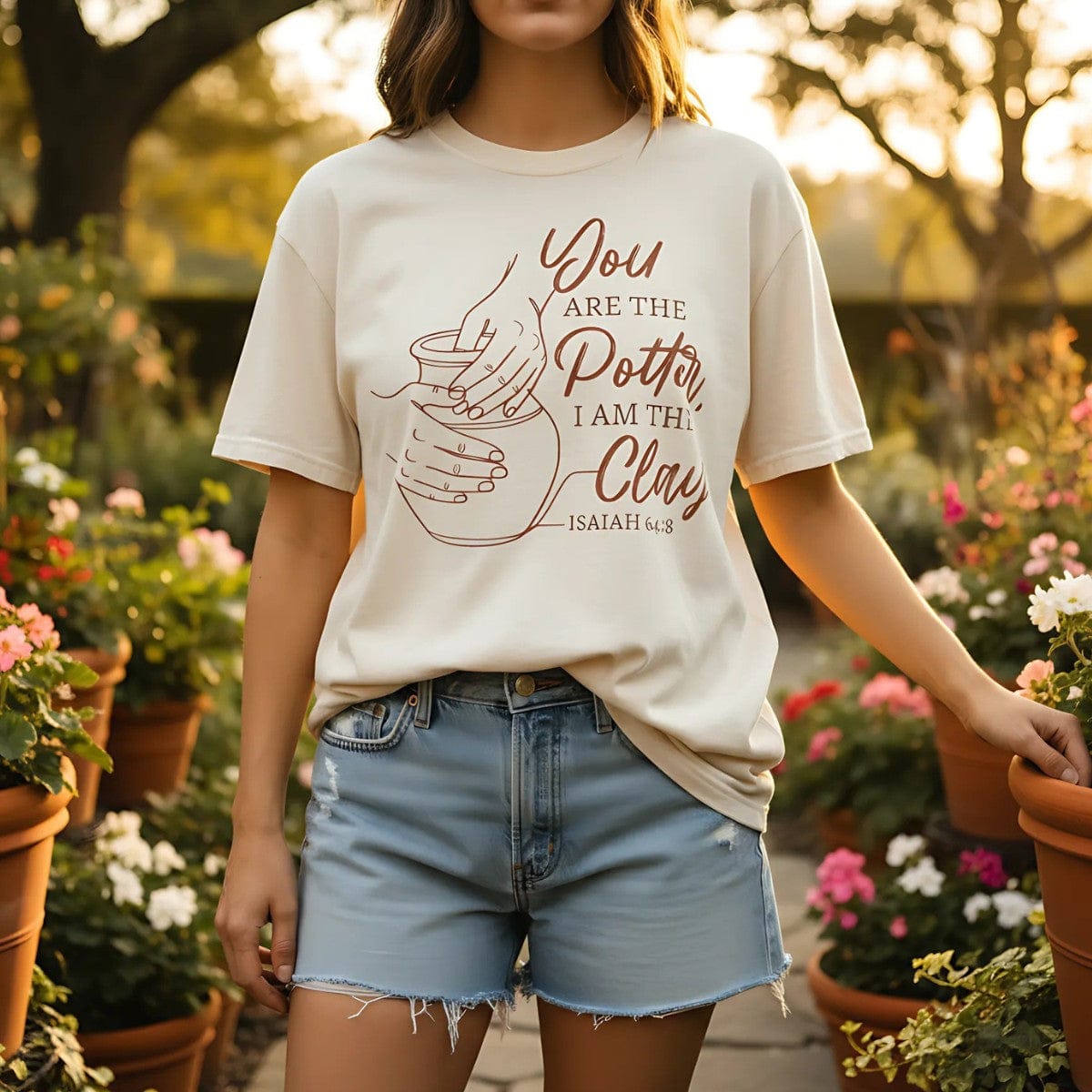 Woman wearing an Ivory Comfort Colors Christian t-shirt with Isaiah 64:8 “You are the Potter, I am the Clay” and pottery line art design. Styled outdoors in a garden setting with flowers, paired with denim shorts for a casual faith-inspired look.