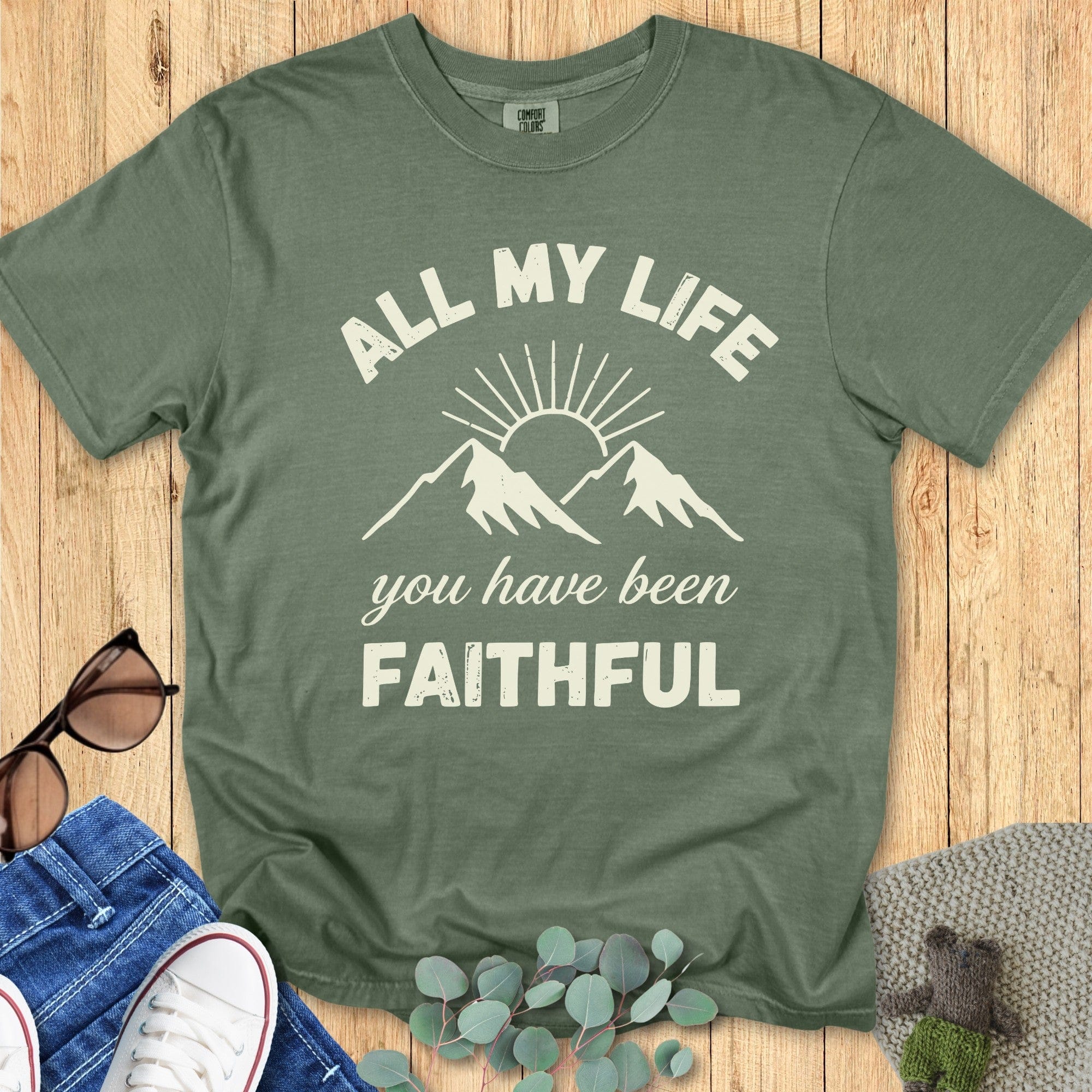 Comfort Colors 1717 moss green Christian T-shirt with mountain and sunrise graphic. Text reads 'All My Life You Have Been Faithful.' Styled flat lay with jeans, white sneakers, sunglasses, and greenery on wood background.
